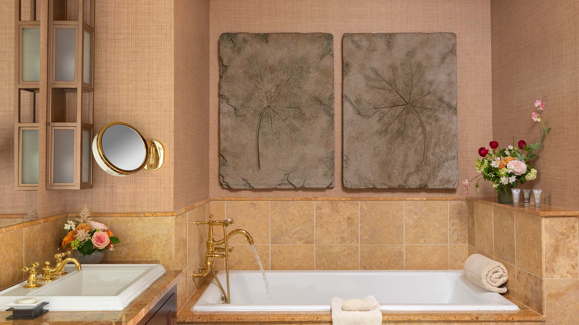 interior shot of the bathroom. There is neutral beige tiling and a white bathtub with gold hardware. There is a sink next to the bathtub. There are flower arrangements placed around the room