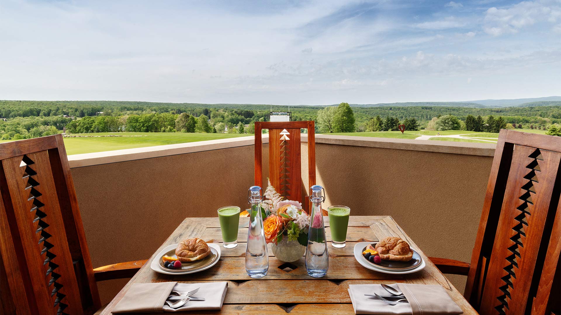 exterior shot of the balcony overlooking the resort grounds. There is a long table on the balcony with wooden chairs lining it. On top of the table is a elegant breakfast spread of croissants, fruit and smoothies