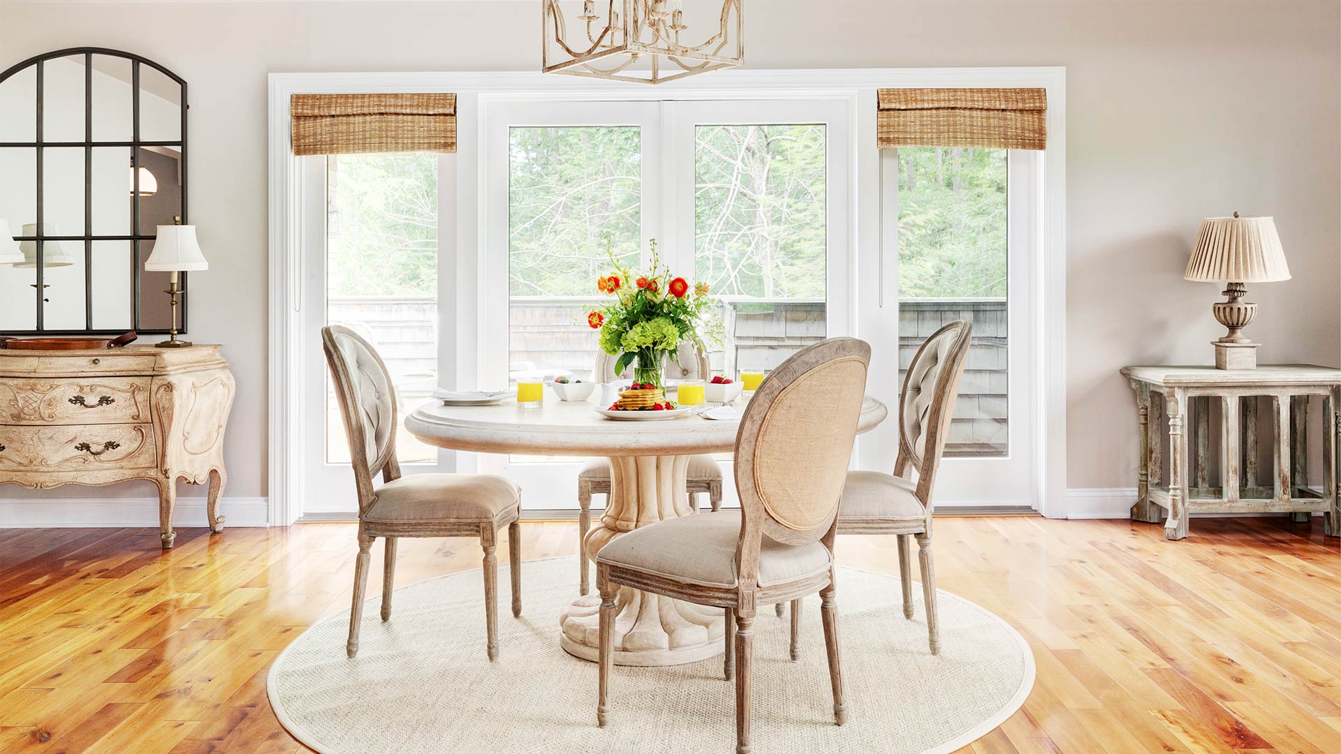 an interior shot of garshak's dining area. There is a round table surrounded by elegant chairs. There is a dresser with a mirror on top to the left of the table and a tab'e with a lamp to the right. There are glass doors behind the dining table that open to the yard.