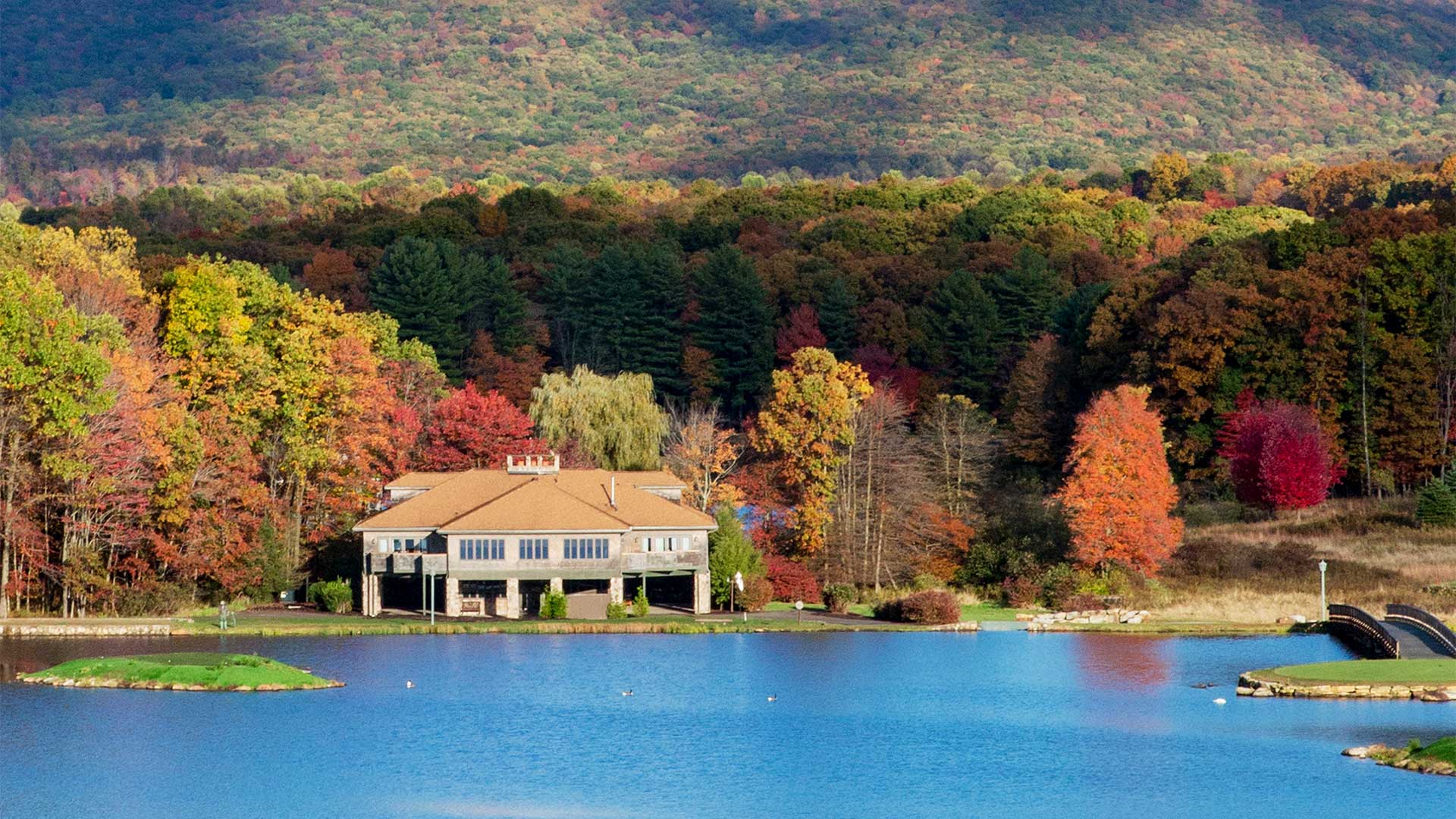 exterior shot of garshak. The house is lakefront. The beautiful fall foliage stretches out in the background and surrounds the house.
