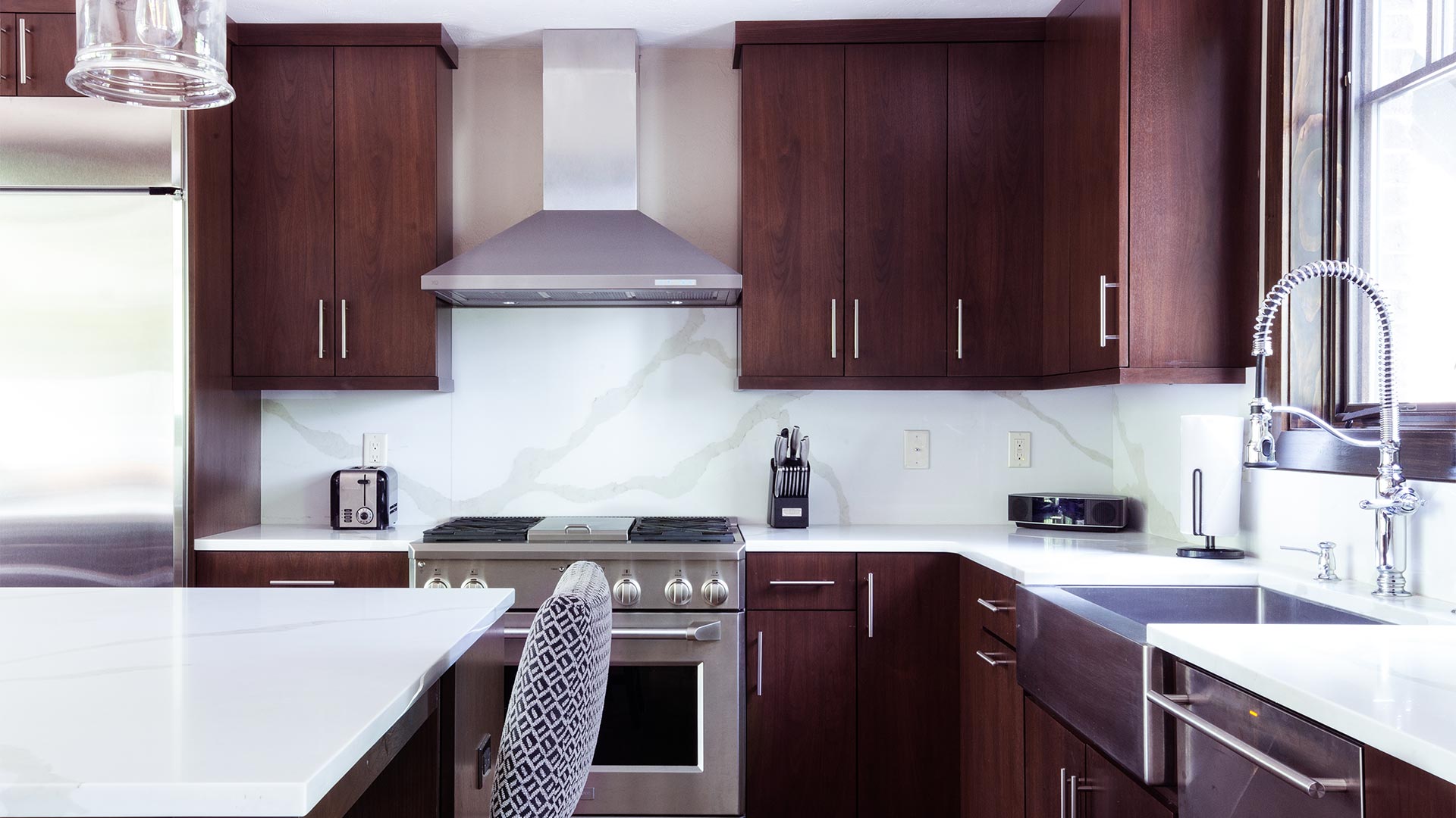 interior shot if the kitchen. There are dark wood cabinets and stainless steal appliances. There are white countertops and a island with chairs.