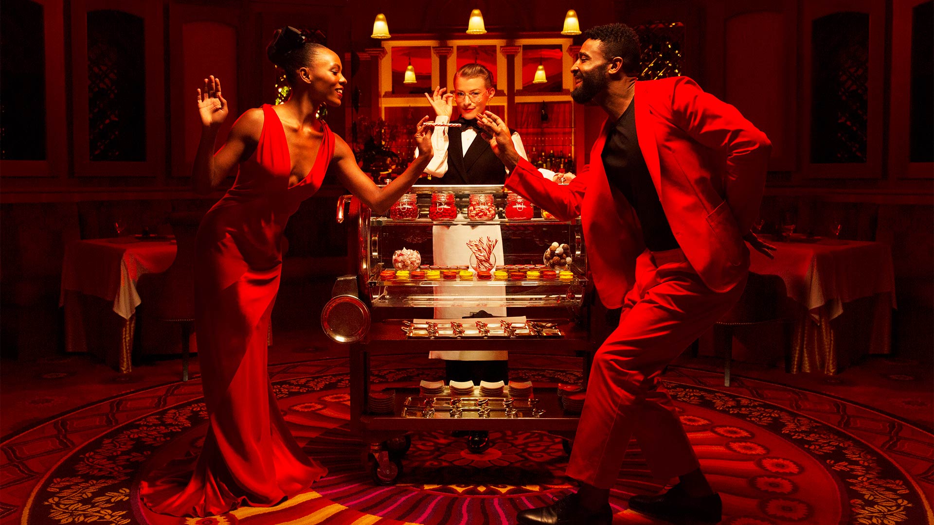 a man and woman in red dancing in front of a waitress standing behind a cart of food