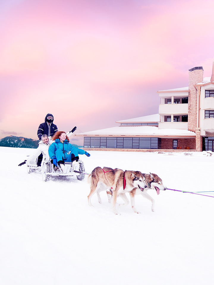 a red haired lady riding a sled that is being pulled by dogs with a hotel in the background