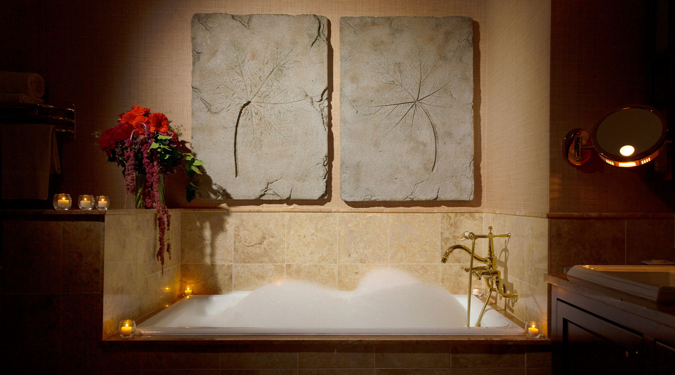 A tile-surround bathtub filled with bubbles with gold fixtures, lit candles, florals, and wall decor in the scene