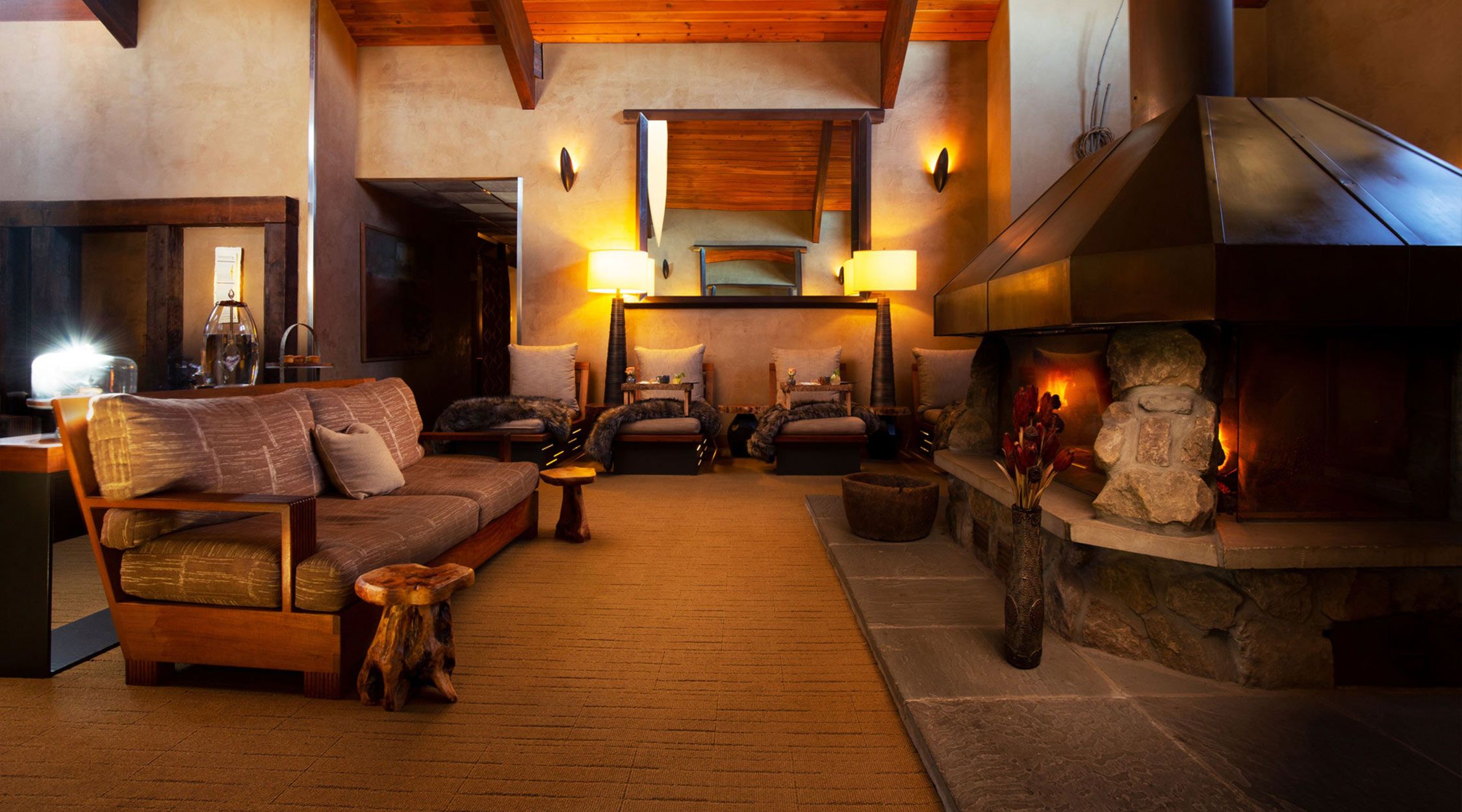 A serene space with high wooden ceilings and natural colors showing a sofa, lounge chairs with large pillows, and a fireplace with an overhead flue vent at the Woodlands Spa at Nemacolin resort