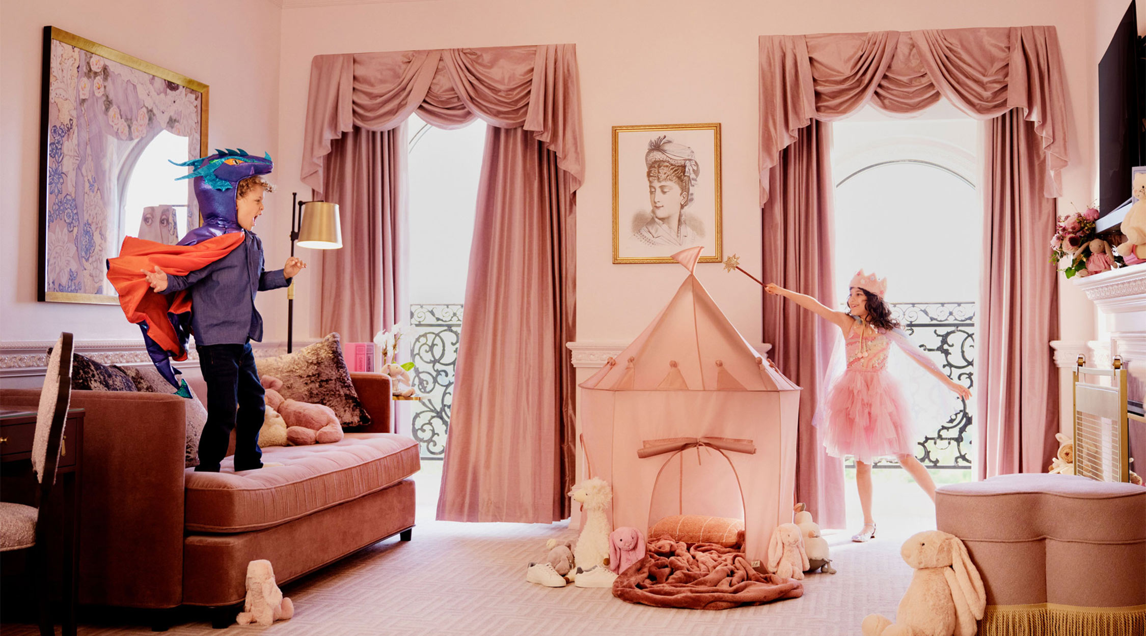 A young boy standing on a pink sofa dressed in a dragon costume watching a young girl dressed as a princess in pink clothes wave a wand over a small, pink castle tent within a pink room with tall windows, tall ceilings, and pinks walls and draperies in a hotel room at The Chateau at Nemacolin resort