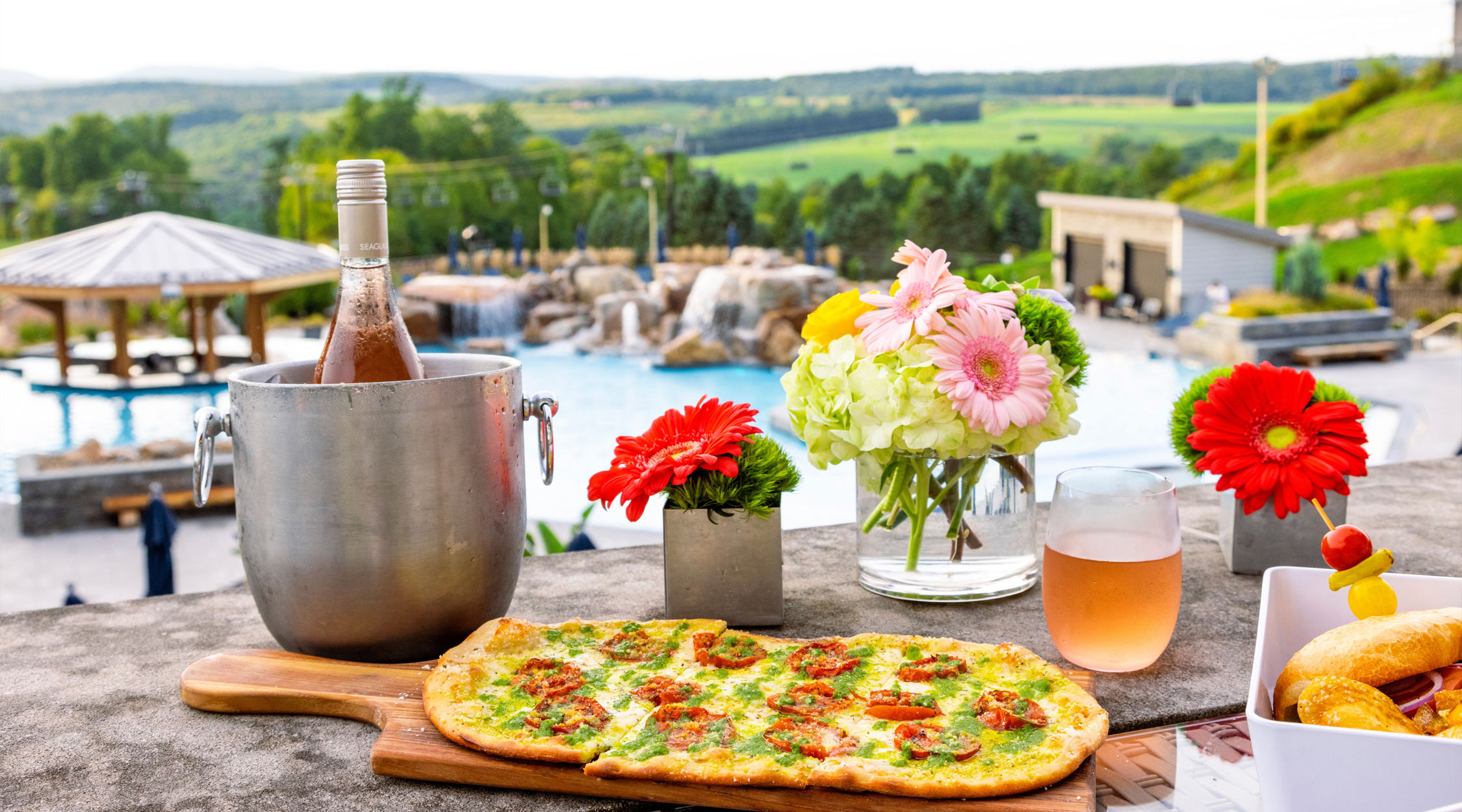 A bottle of rose wine in a silver ice bucket behind a flatbread pizza and summer flowers on a table overlooking a pool and the mountains