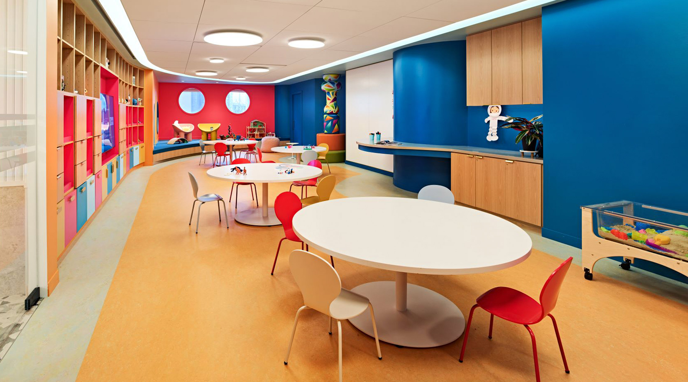 a colorful room containing two, round white tables and red and white chairs with a blue wall opposite a wall of bookshelves, and a red wall with two round windows at the far end