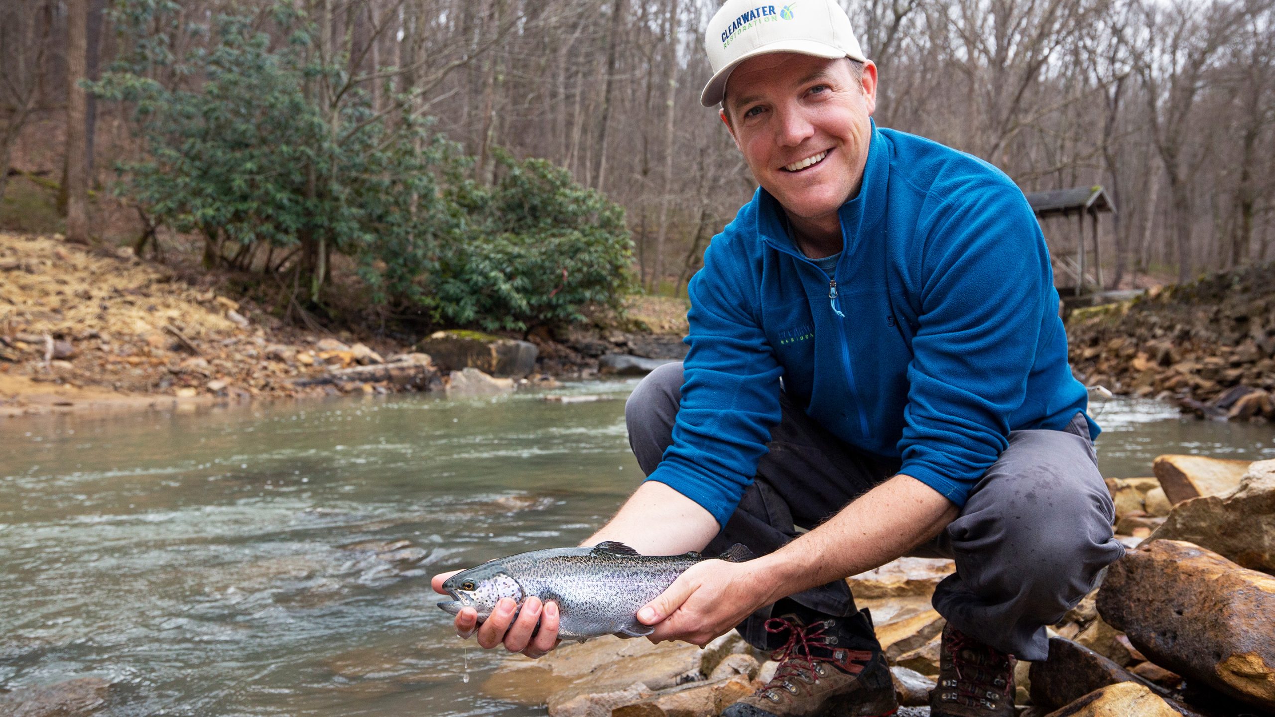 Image of a man holding a trout next to a fishing stream in the forest