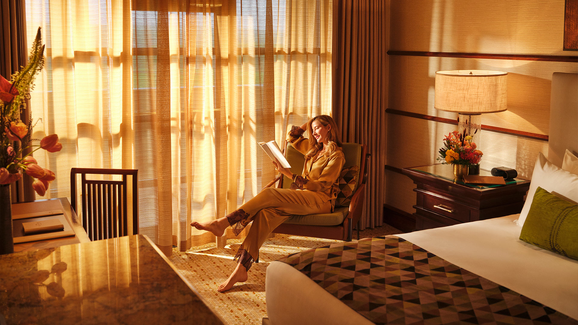 A woman seated on a comfortable chair while reading a book in a room while a golden glow streams through sheer floor-to-ceiling curtains. In the foreground a bed and desk with chair are seen.