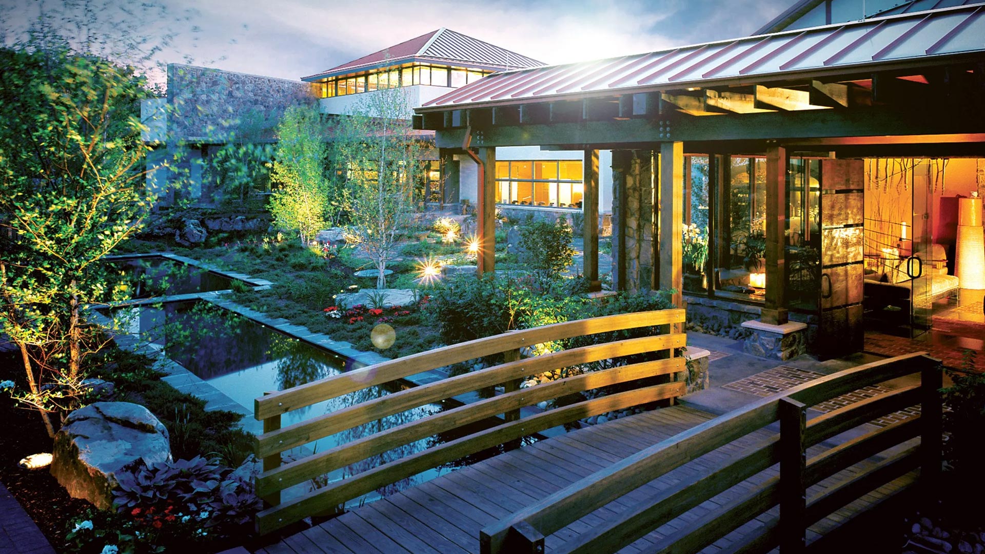 A wooden bridge leading to the illuminated modern, glass Woodlands Spa at Nemacolin building with a rectangular pond of water flowing under the bridge