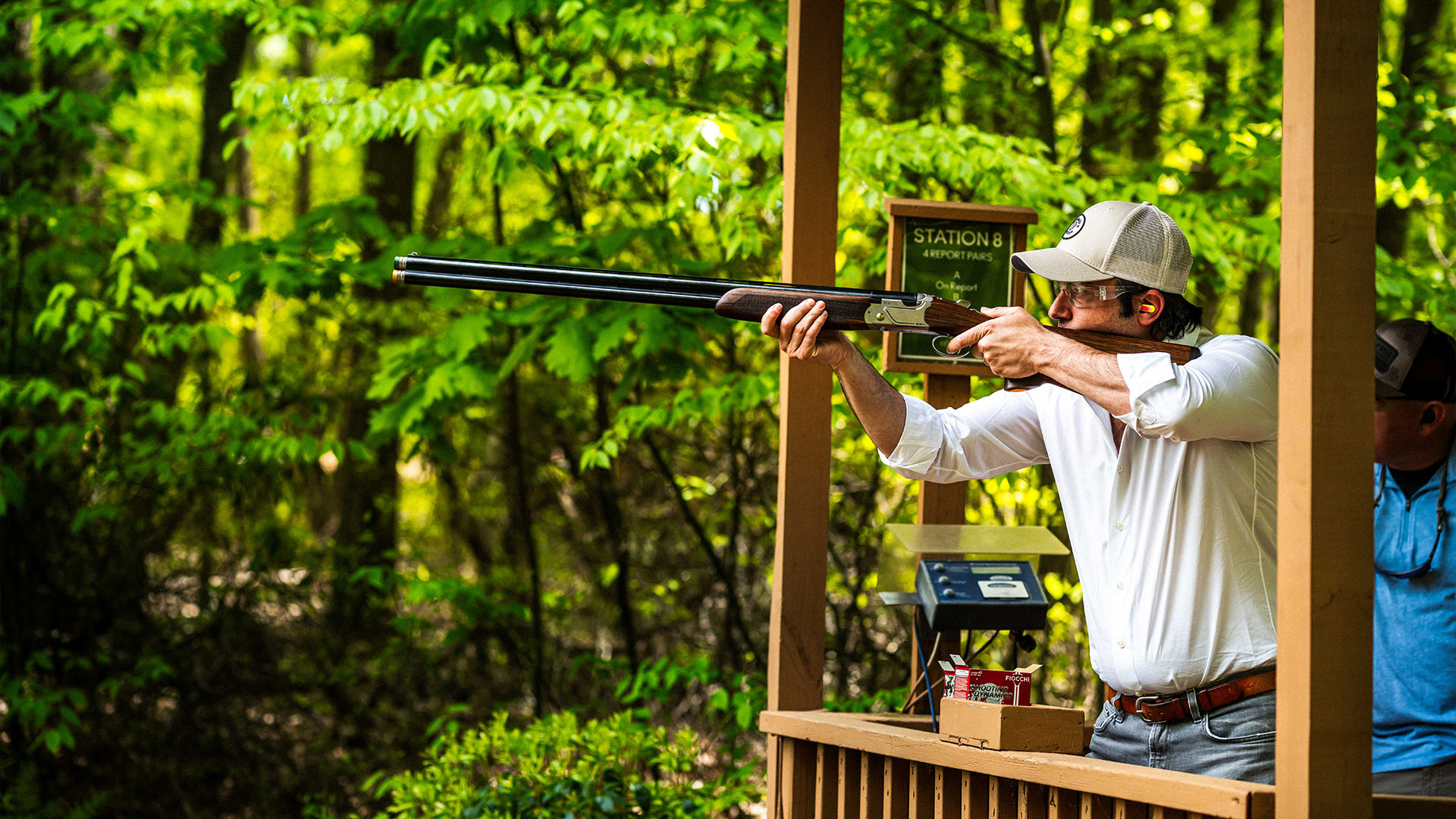 Amidst a forest of green trees, a man in a white shirt, white hat, and blue jeans holds a rifle up as he is in the act of aiming as he stands on a wooden stand between two wooden columns with a railing in front of him with a box of shotgun shells on the railing