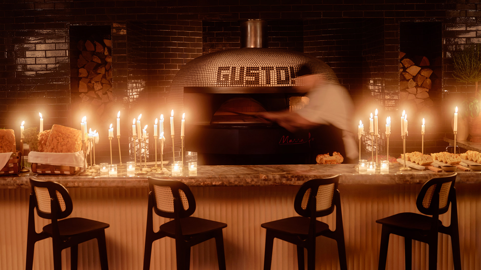 A large pizza oven with a man in a white chef's coat and black hat putting in a pizza. Surrounding the oven are dozens of lit candles, and in front of the oven is a bar with four bar chairs.