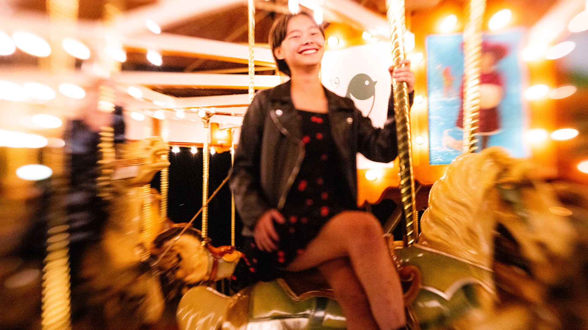 A woman with short brown hair, wearing black leather jacket over a black dress dotted with red is smiling as she sits side-saddle and cross-legged on a carousel horse while the carousel was blurred in the background