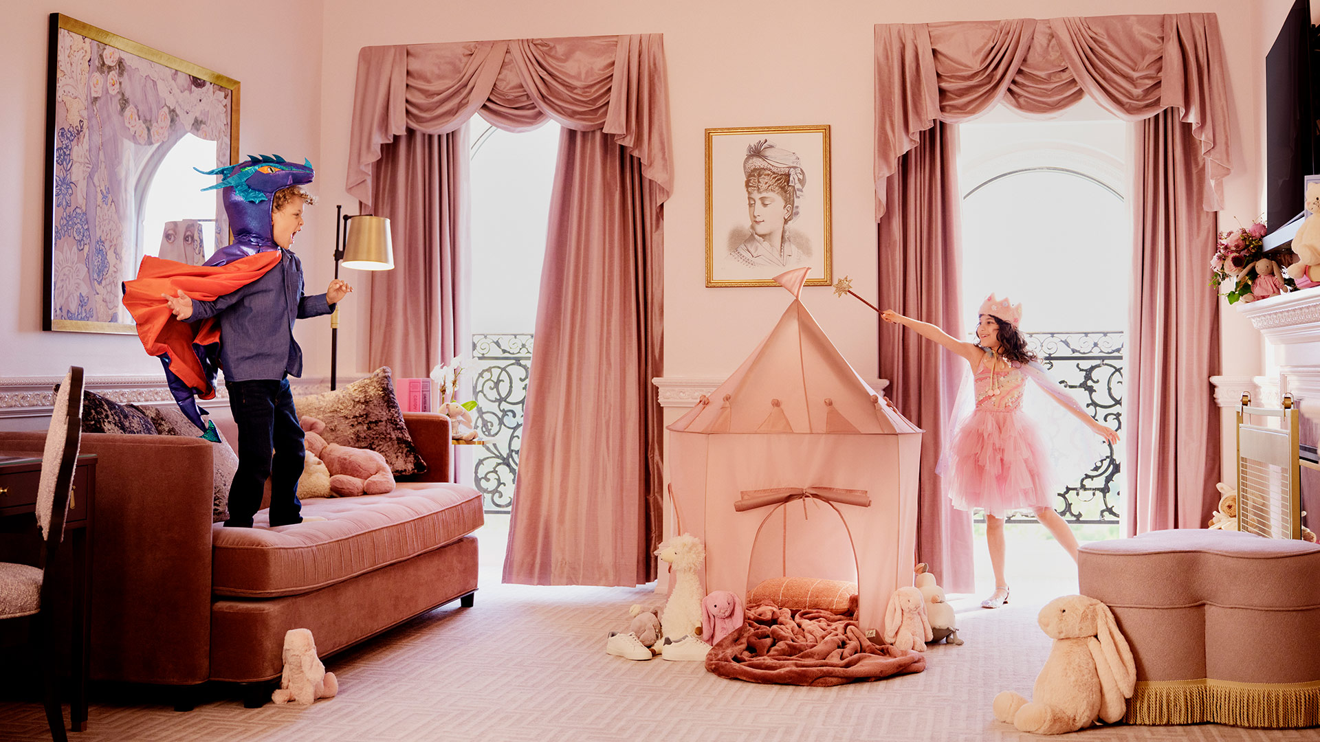 A pink room at The Chateau at Nemacolin with a boy dressed as a dragon or dinosaur is jumping on a rose-colored sofa while a girl stands before floor-to-ceiling windows dressed as a priness in a pink gown and crown waving a wand over a pink tent set up in the room