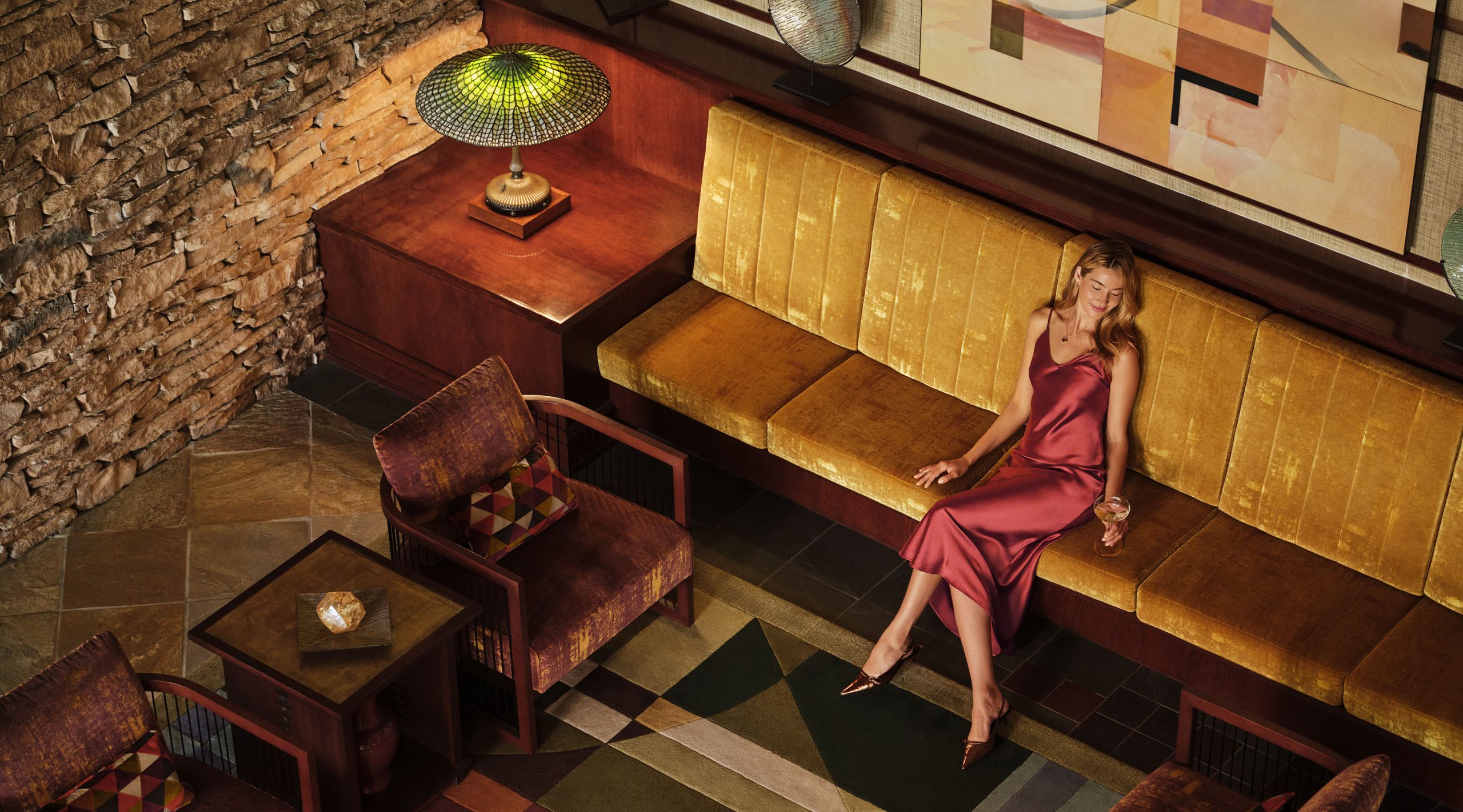 Elegant guest relaxing with a cocktail in the sophisticated lobby at Nemacolin resort's Falling Rock boutique hotel, featuring warm lighting, golden velvet seating, and refined mid-century design elements.