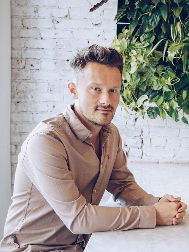 A man sitting a a whilte table in a room surrounded by white bricks and a hanging plant